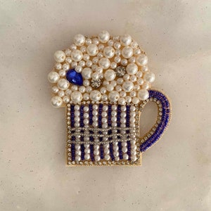 May include: A white and blue beaded beer stein with a white pearl foam top. The stein has a blue handle and is decorated with small white and blue beads.