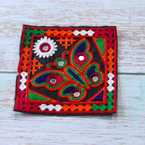 Puede incluir: Un parche cuadrado bordado de colores con un diseño de mariposa. La mariposa está hecha de hilo rojo, verde, azul y blanco y tiene pequeños espejos para los ojos. El parche está rodeado por un borde de hilo rojo, naranja, blanco y verde.