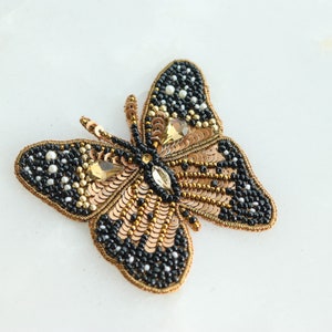 Puede incluir: Un broche de mariposa con cuentas negras, doradas y bronce. El diseño presenta lentejuelas y abalorios, con cristales transparentes y ámbar. Las alas están delineadas con hilo dorado, creando un efecto texturizado.