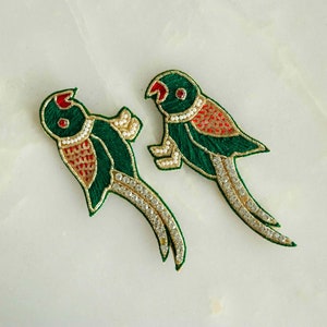 May include: Two embroidered parrot patches. The parrots are green with red and gold accents, and embellished with white beads and clear rhinestones. The patches are on a white marble surface.