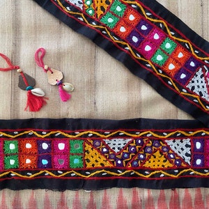 May include: Two black fabric trims with colorful embroidery and mirror embellishments. The trim is decorated with geometric patterns and small mirrors. The trim is perfect for adding a touch of ethnic flair to any project.