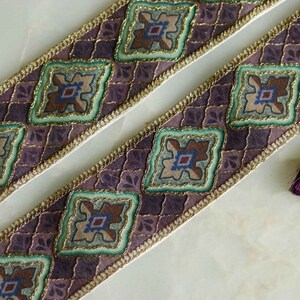 May include: Two decorative ribbons with a repeating square pattern in shades of purple, teal, and gold. The ribbons have a gold trim and are accompanied by two small tassels, one purple and one teal.