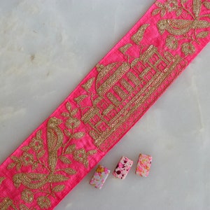 以下が含まれることがあります： ピンクの生地のトリムに、鳥、花、鳥かごのデザインの金色の刺繍が施されています。