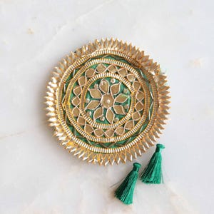 Puede incluir: Un objeto decorativo redondo de color dorado con un centro verde y detalles intrincados. Dos pequeñas borlas verdes están colocadas debajo. El diseño presenta un patrón floral y un acabado metálico texturizado.
