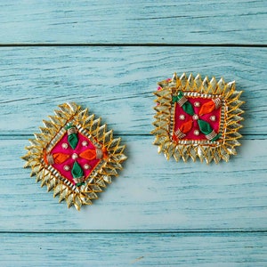 Puede incluir: Dos adornos decorativos dorados, rosas, verdes y naranjas con un diseño de estrella. Los adornos están sobre una superficie de madera azul.