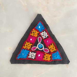 Peut inclure: Un patch triangulaire en tissu avec des broderies colorées et des accents miroir. Le patch présente un motif géométrique avec des motifs floraux dans des tons de bleu, rose, jaune et blanc.