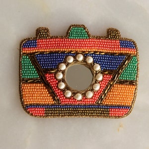 May include: A colorful beaded mirror with a geometric design in shades of orange, red, blue, and green. The mirror is surrounded by a circle of white pearls.