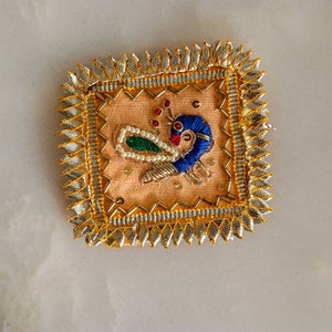May include: A square-shaped embroidered patch with a peacock design. The peacock is blue, green, and white with a gold outline. The patch is surrounded by a gold and silver border.
