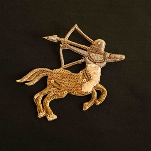 Könnte beinhalten: Gold gestickter Aufnäher eines Zentauren mit Bogen und Pfeil. Der Zentaur blickt nach rechts und hat einen silberfarbenen Körper.