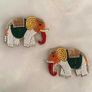 Puede incluir: Dos adornos de elefante de cuentas con cuentas blancas, verdes, naranjas y rojas. Los elefantes miran hacia la derecha y tienen una silla de montar en la espalda.
