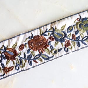 Peut inclure: Garniture en tissu blanc avec des motifs floraux brodés dans des tons de brun, vert et bleu. Les fleurs sont stylisées et ont un aspect vintage.