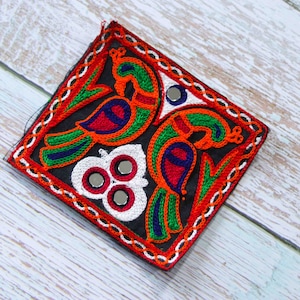 Puede incluir: Un parche cuadrado de tela negra con bordado colorido de dos pájaros y un diseño floral. Los pájaros son verdes, naranjas, rojos y azules. El diseño floral es blanco con tres pequeños espejos redondos en el centro.