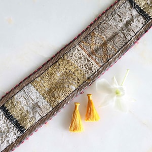 Pode incluir: Uma faixa têxtil decorativa com um design de patchwork com renda e contas douradas, brancas e bege. As bordas são aparadas com uma borda marrom e costura rosa. Também estão presentes dois pingentes amarelos e uma flor branca.