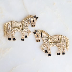 Puede incluir: Dos parches con forma de caballo elaborados con intrincados abalorios. Cada caballo presenta una paleta de colores oro metálico y crema con cuentas negras que detallan los cascos, los ojos y la crin. Incluye un patrón de silla de montar.