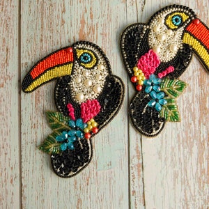 May include: Two beaded toucan patches. The toucans have black bodies, orange and yellow beaks, and are embellished with pearls, pink and blue floral accents, and green leaves. The patches are on a weathered, light-colored wooden surface.
