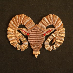 May include: A gold embroidered ram's head with red eyes. The ram's head is a decorative piece that can be used for a variety of purposes, such as a patch or an embellishment.