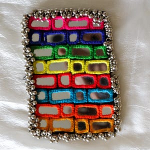 Puede incluir: Un parche rectangular que presenta una cuadrícula de pequeños espejos enmarcados por hilo de ganchillo de colores vibrantes en rosa, azul, verde, amarillo, rojo y naranja. El borde está adornado con una hilera densa de cuentas metálicas plateadas.