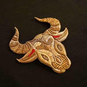 Könnte beinhalten: Gold gestickter Bullenkopf-Patch mit aufwendigen Details und roten Akzenten auf schwarzem Hintergrund. Der Patch zeigt ein detailliertes Design des Kopfes, der Hörner und der Gesichtszüge des Bullen. Dieser dekorative Patch eignet sich für Kleidung oder Accessoires.