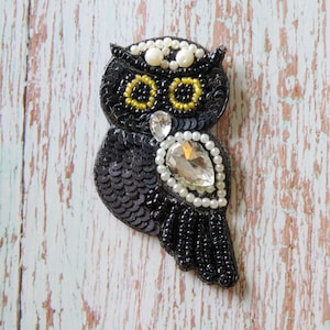 May include: An owl-shaped brooch crafted with black sequins, black beads, and white pearls. The eyes are outlined in yellow beads, and the chest features a large, clear, teardrop-shaped crystal surrounded by a border of small white pearls.