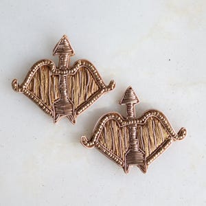 Puede incluir: Dos colgantes artesanales de metal color cobre con forma de arco y flecha. Cada colgante presenta un arco detallado con una flecha que apunta hacia arriba. Los colgantes son probablemente para la fabricación de joyas o manualidades.