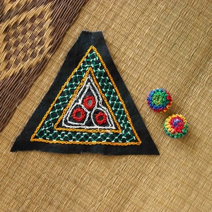 Puede incluir: Un parche triangular de tela negra con bordados coloridos, que presenta un borde verde y amarillo, un triángulo interior blanco y tres círculos rojos. También se muestran dos pequeños botones redondos de ganchillo de colores.