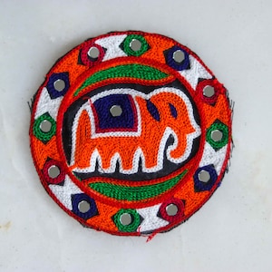Puede incluir: Un parche redondo bordado de colores vivos con un diseño de elefante naranja. El parche está decorado con espejos y tiene un fondo negro.