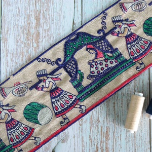 May include: A beige fabric trim with a repeating pattern of figures playing musical instruments and carrying a palanquin. The figures are embroidered in blue, red, green, and white.