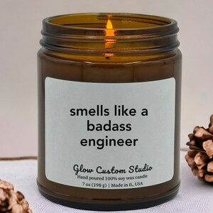 May include: A brown glass candle jar with a white label that reads "smells like a badass engineer". The label also includes the text "Glow Custom Studio", "Hand poured 100% soy wax candle", "7 oz (198 g)", and "Made in IL, USA".