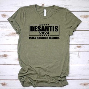 May include: A green t-shirt with black text that reads "DESANTIS 2024 MAKE AMERICA FLORIDA".