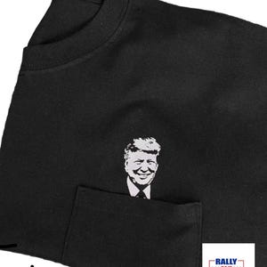May include: Black t-shirt with a pocket featuring a white silhouette of a man's face. The text "RALLY 24/7 POINT" is printed on a red, white, and blue banner in the bottom right corner.