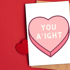 May include: A white card with a pink heart-shaped candy with the text "YOU A'IGHT" in white.