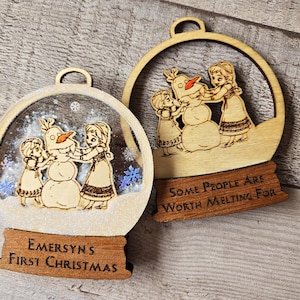 May include: Two wooden Christmas ornaments featuring a snowman and two girls. The ornament on the left says "Emersyn's First Christmas". The ornament on the right says "Some People Are Worth Melting For".