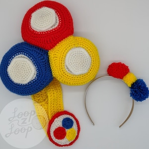 Puede incluir: Una diadema de crochet con tres pompones de colores y un juego a juego de tres bolas de crochet. Las bolas son rojas, amarillas y azules con centros blancos. La diadema es plateada y los pompones son rojos, amarillos y azules. El texto "Loop Z Loop" es visible en la imagen.