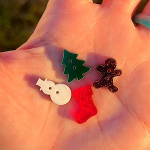 May include: A collection of Christmas-themed resin charms, including a green Christmas tree, a white snowman, a red stocking, and a brown gingerbread man with glitter. The charms are small and colorful, perfect for crafting or jewelry making.