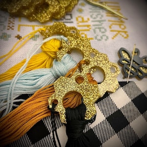 May include: Gold glitter thread organizers with tassels in blue, yellow, and orange. The organizers are on a black and white checkered fabric with a bee cutout.