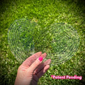 May include: A clear, heart-shaped quilting template with concentric circular markings and measurements. The template is held against a backdrop of green grass. The text "*Patent Pending" is visible in the lower right corner.