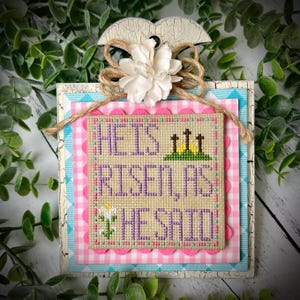 May include: A cross stitch design on a wooden plaque with a pink and blue checkered background. The design reads "He is risen, as He said".