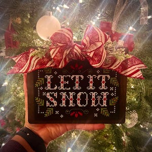 May include: A wooden sign with a red and white striped bow, decorated with green sprigs. The sign reads "Let It Snow!" in white embroidery.