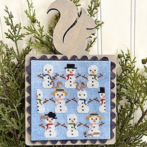 May include: A cross-stitch winter scene featuring multiple snowmen with scarves and hats on a light blue background. The cross-stitch is framed with a scalloped edge and a wooden squirrel topper. The piece is surrounded by evergreen branches.