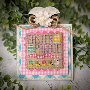 May include: A cross-stitched Easter-themed wall hanging with a pink and white gingham background. The cross-stitch design features the text "Easter Parade" with a banner that reads "Hop Hop Hop" and a bunny rabbit with chicks.