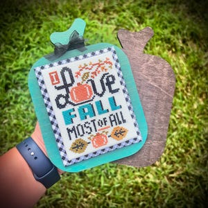 Pode incluir: Uma placa de madeira com uma borda verde-azulado e um design bordado em ponto cruz que diz "I Love Fall Most of All". A placa apresenta abóboras e folhagens de outono. Uma fita preta está amarrada no topo.