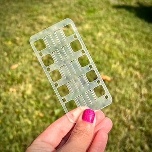 Puede incluir: Una plantilla de plástico transparente con una cuadrícula de formas rectangulares y cuadradas. La plantilla se sostiene en una mano con esmalte de uñas rosa.