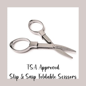 May include: A pair of open, silver-colored, foldable scissors. The scissors have two finger holes and a blade. The text "TSA Approved Slip & Snip Foldable Scissors" is below the scissors.
