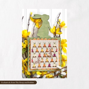 May include: A cross-stitch pattern of a bunny surrounded by carrots and other vegetables, framed in a green wooden frame with a scalloped edge. The frame is decorated with yellow flowers.