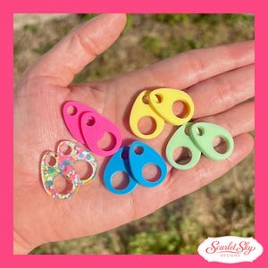 May include: A collection of colorful stitch markers in various shapes and colors, including pink, yellow, blue, and green. Some markers have a glittery, multi-colored design. The markers are held in a hand.