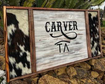 Op de afbeelding: Een rustiek houten bord met een witgeschilderde achtergrond en een bruine rand. Het bord heeft de tekst "CARVER TA" in zwarte letters met een decoratieve krul. Het bord is omlijst met een bruin houten frame en heeft een rand van koeienhuid.