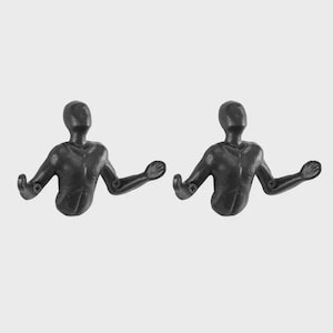 May include: Two black metal wall hooks shaped like human figures with outstretched arms. Each hook has a screw for mounting. The hooks are designed to hold items and add a decorative touch to any space.