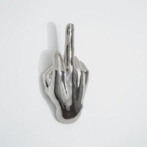 May include: A silver-colored hand sculpture with the middle finger extended upwards. The sculpture is highly reflective and appears to be made of metal. The hand is mounted on a white surface, casting a shadow.