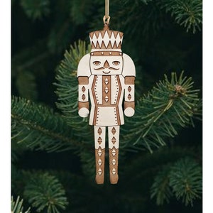 Puede incluir: Un adorno de madera blanco en forma de cascanueces con detalles marrones, colgado de un árbol de Navidad verde. El cascanueces tiene una corona, bigote y un diseño estampado en su uniforme.
