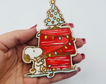 May include: A wooden Christmas bauble featuring Snoopy standing in front of his red doghouse decorated with Christmas lights. A small Christmas tree sits on top of the doghouse.